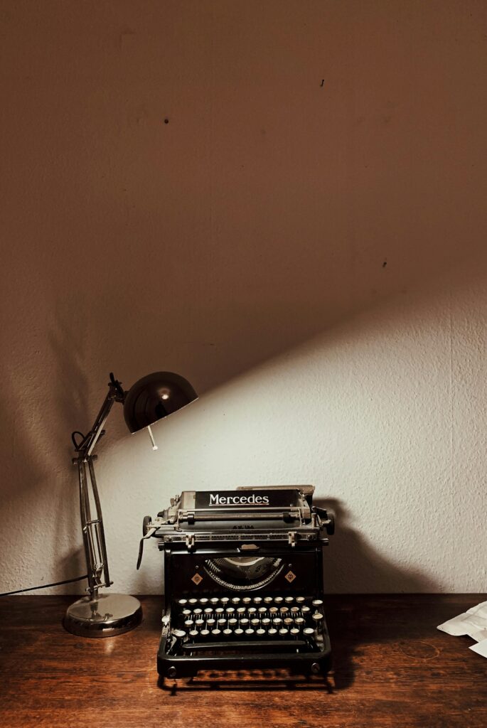 A black typewriter sits on a wooden desk, with a lamp shining a light over it.